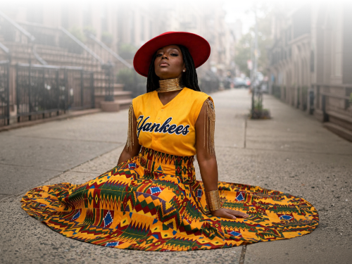 Linda Orji seated in her Yankees jersey and flowing Kente skirt — original portrait in a concrete setting