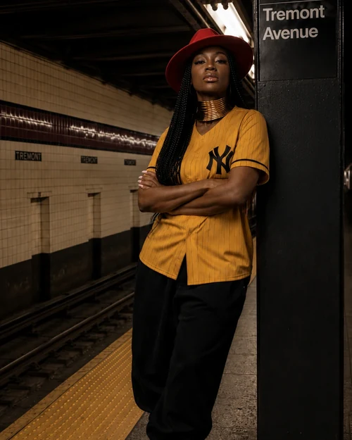 Linda Orji in her mustard New York jersey and red wide-brim hat standing confidently on a New York City subway platform